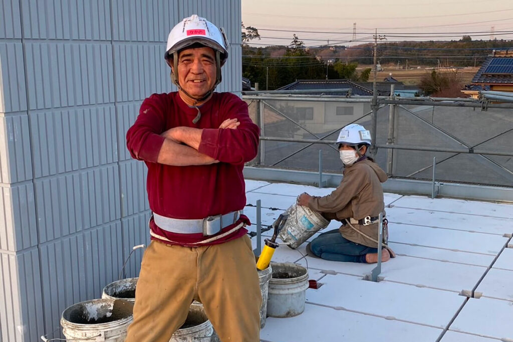 現場で腕を組んで立つベテラン作業員と、目地工事中の作業員